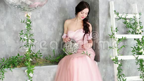 穿着粉红色长裙的迷人女士坐在植物工作室装饰的灰色角落里视频的预览图