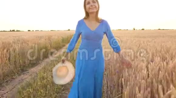 日落时分穿着蓝色连衣裙和帽子的漂亮女人穿过麦田自由概念日落时的麦田视频的预览图