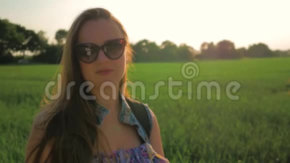 一个戴眼镜的年轻漂亮女孩的肖像她的长发在日落时站在田野里视频的预览图