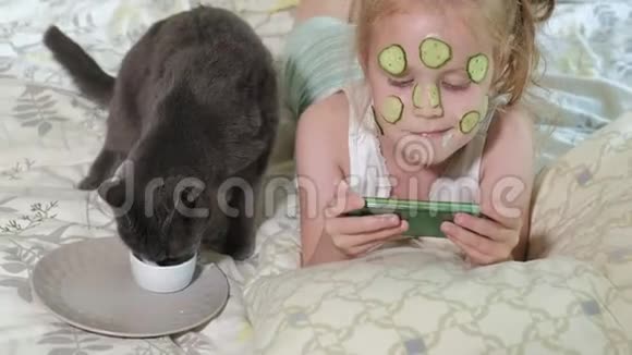 一个漂亮的小女孩兴高采烈地在家里做黄瓜面具享受猫坐在附近的智能手机视频的预览图