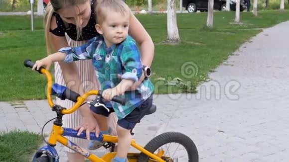 年轻女子在公园教她蹒跚学步的儿子骑自行车的慢镜头视频的预览图