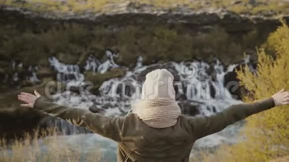 年轻的旅行女性举起双手欣赏冰岛美丽的瀑布美景视频的预览图