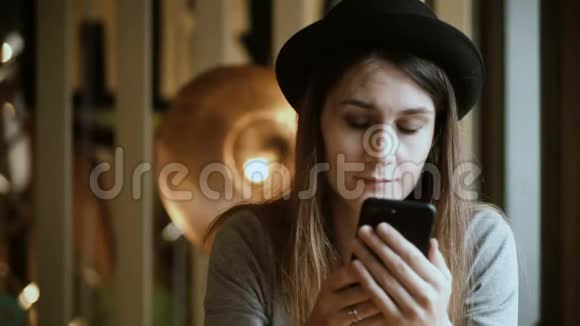 肖像如果美丽的女人坐在咖啡馆和使用智能手机有魅力的戴帽子的女人从手机上聊天视频的预览图