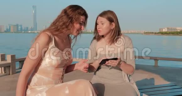 两个穿着浅色连衣裙的微笑女孩正在智能手机上看照片背景为蓝水湖和城市景观视频的预览图