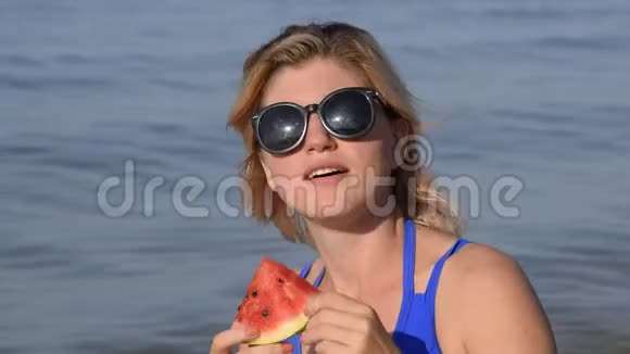 一个戴太阳镜的女人坐在海滩上吃西瓜一个成熟的西瓜在金发女郎手中视频的预览图