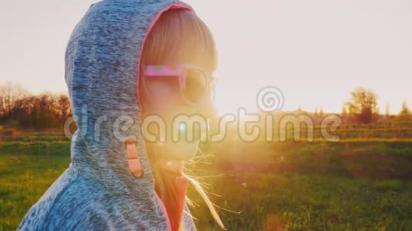 六岁的女孩穿着带帽的运动服站在夕阳的背景下戴着太阳镜玫瑰色的眼镜视频的预览图