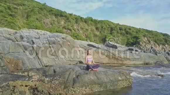 女孩坐在瑜伽位置海浪拍打岩石视频的预览图