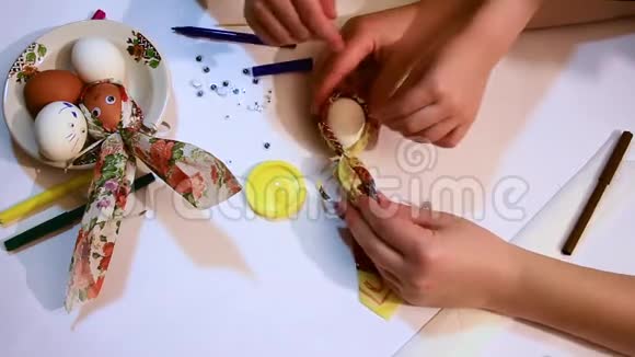 一个女孩和一个孩子用一张彩色餐巾和一条闪亮的丝带装饰一个白色的鸡蛋并画出有趣的图案视频的预览图