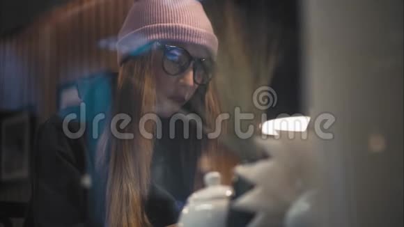 一位时髦的女性戴着时尚的眼镜和帽子坐在咖啡馆里用笔记本电脑工作从窗口观看视频的预览图