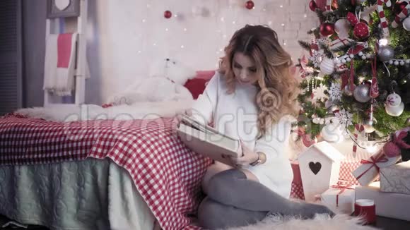 年轻的美女在圣诞树附近的床上看着相册视频的预览图
