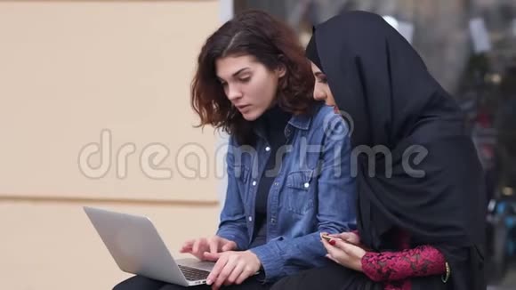 两个女朋友坐在外面用笔记本电脑跨越文化友谊年轻的穆斯林女人戴着黑色头巾视频的预览图