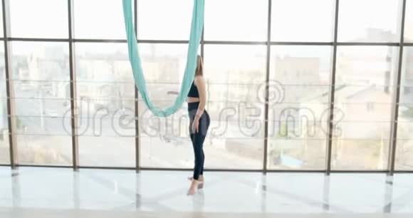 漂亮的年轻女人在有大窗户的房间里做飞行瑜伽运动有魅力的女性在她的身体里工作视频的预览图