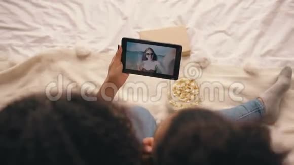 两个女朋友坐在床上在家里和他们的朋友度假在平板电脑上用Skype聊天视频的预览图