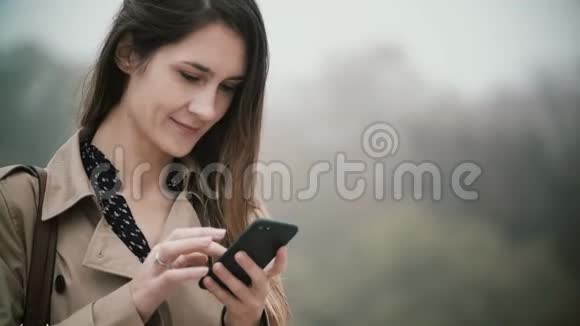 雾蒙蒙的公园里年轻美女的肖像微笑的女性在早上使用智能手机视频的预览图