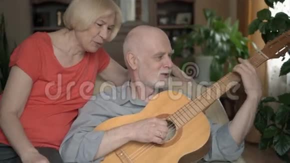 家里的老夫妇丈夫弹吉他妻子坐在扶手椅上唱歌和微笑视频的预览图