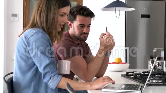 漂亮可爱的年轻夫妇用他们的笔记本电脑在家里的厨房里吃早餐视频的预览图