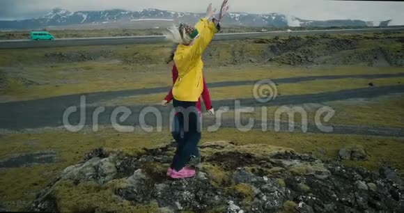 鸟瞰两位身穿鲜艳衣服的幸福女子在山顶上翩翩起舞在冰岛的熔岩领域旅游视频的预览图
