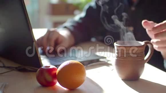 一个男人在用笔记本电脑他喝热茶悬崖上躺着苹果和橘子健康的饮食专注于杯子视频的预览图