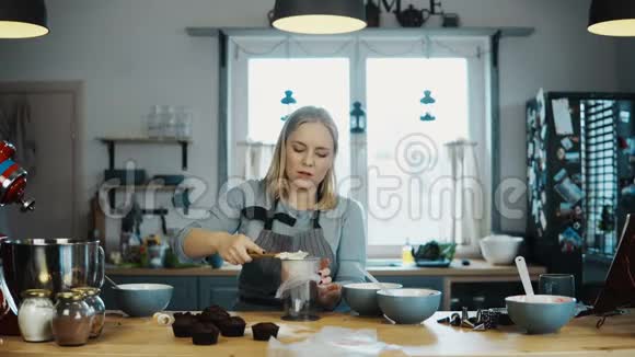 年轻漂亮的女人站在厨房里做纸杯蛋糕金发女郎把奶油放进糕点袋里视频的预览图