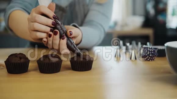 女性手的特写镜头将糕点袋中的奶油放在巧克力纸杯蛋糕上做松饼的年轻女人视频的预览图