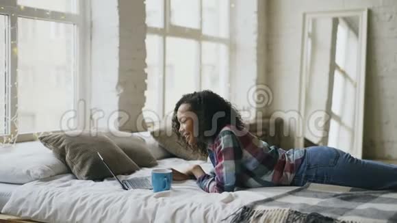 漂亮的混血少女使用笔记本电脑清洗分享躺在家里床上的社交媒体视频的预览图