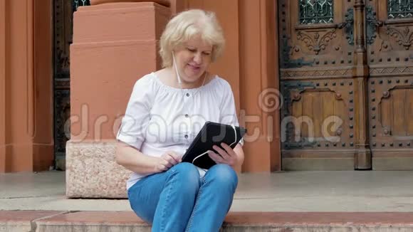 老年妇女坐在台阶上使用带有耳机的平板电脑背景中的老式建筑视频的预览图