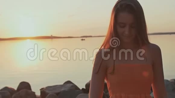 一个穿着橙色连衣裙的年轻女孩在海滩上的日落时摆出姿势玩她的头发视频的预览图