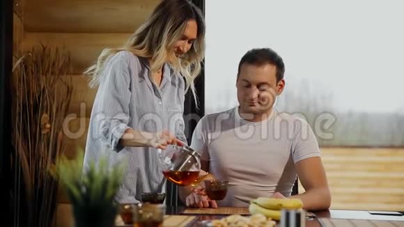 穿着睡衣的漂亮女人倒了她丈夫的茶在厨房里拥抱他喝茶情人们视频的预览图