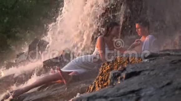 一个穿着白色连衣裙长发的漂亮女孩和一个男人躺在瀑布下的岩石中抚摸着他的脸视频的预览图