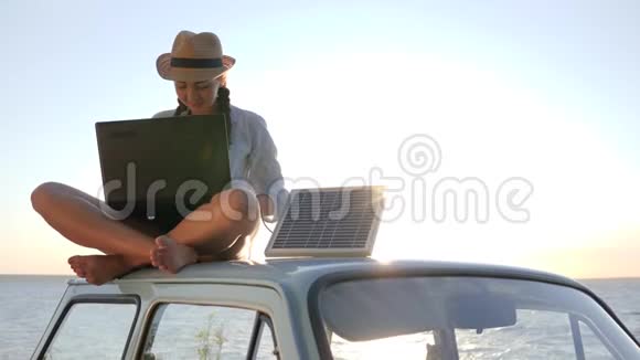 年轻女性在阳光下坐在老式汽车上的背景海滩上利用太阳能电池驱动电脑旅行视频的预览图