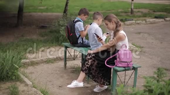 两个男孩和女孩在放学后使用他们的电话可爱的男孩坐在长凳上玩网络游戏视频的预览图