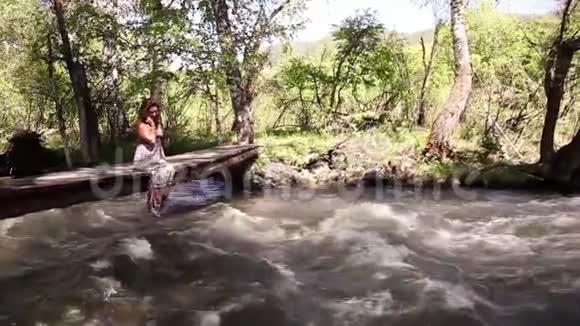 一个美丽的女孩在湖边放松坐在一座木桥的边缘双脚在水面上摆动视频的预览图