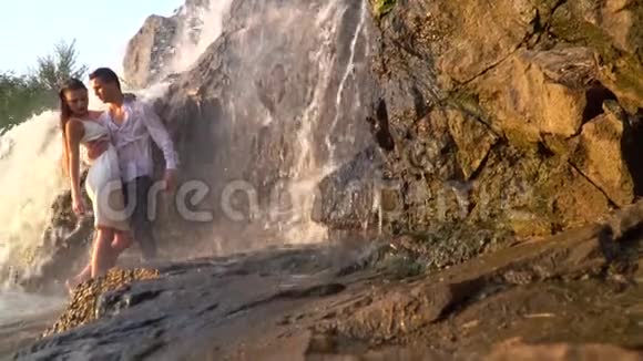 日落时分一个男人抱着一个女孩站在瀑布下的岩石中动作缓慢视频的预览图