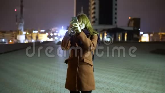 迷人的年轻女性在夜间漫步街头时在智能手机上拍照慢动作视频的预览图