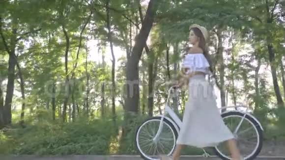 一个漂亮的黑发女人穿着白色裙子一件衬衫在阳光下滚动一辆城市自行车在公园的小路上视频的预览图