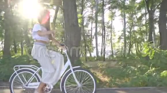 穿着白色裙子的布鲁内特穿着一件衬衫在公园的小路上骑着一辆城市自行车在阳光下骑着摘下一顶草帽视频的预览图