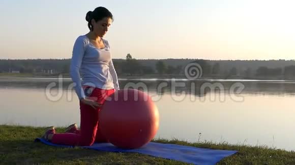 一个运动的女人在日落时膝盖上滚着一个红色的健身球视频的预览图
