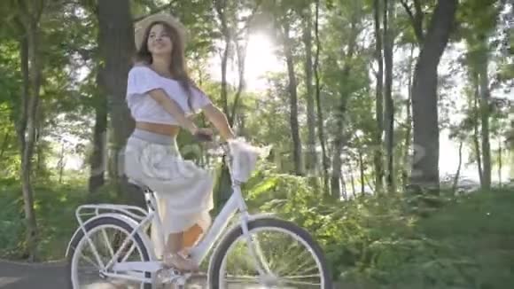 一件衬衫穿着一条白色裙子漂亮的深色头发沿着公园里的小路骑着一辆城市自行车摘下她的草帽和草帽视频的预览图