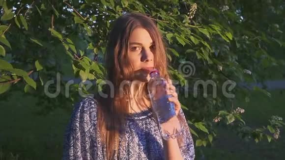 美丽的女性徒步旅行者在日落时在森林里饮水穿裙子的女孩从塑料瓶里喝凉水视频的预览图
