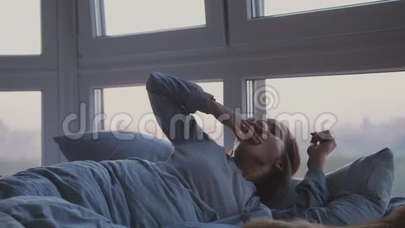 清晨美丽的黑发女人醒来躺在床上打哈欠在日出时在全景窗旁的房间里视频的预览图