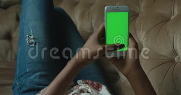 这位美国黑人妇女躺在沙发上用带有绿色屏幕的手机放松和聊天关闭视频的预览图