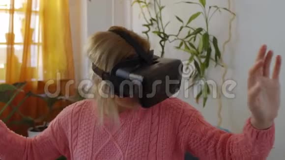 穿着粉红色毛衣的女人在家享受在现代扶手椅中使用虚拟现实护目镜视频的预览图