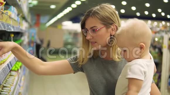 年轻漂亮的母亲戴着眼镜抱着她的孩子一边在挑选婴儿食品视频的预览图