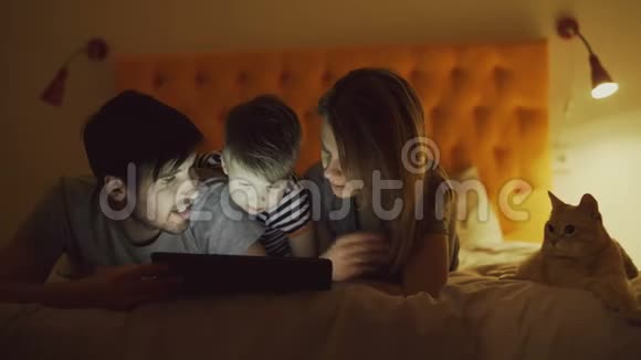 幸福的家庭有一个小儿子和一只有趣的猫躺在家里的床上用平板电脑看卡通电影视频的预览图