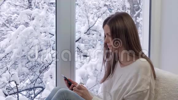 坐在窗台上的漂亮女孩用带耳机的智能手机冬天在外面视频的预览图