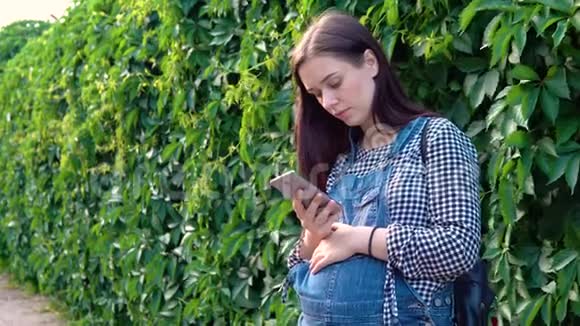 年轻的怀孕女孩穿着工作服看着电话绿色的背景视频的预览图