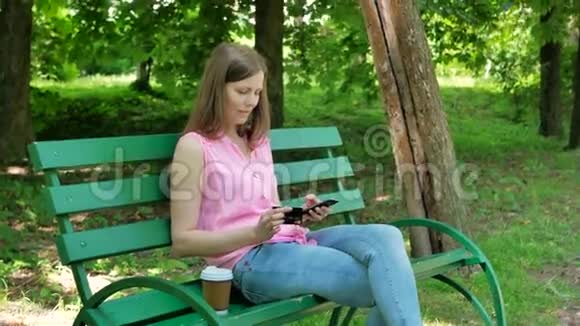 漂亮女孩在公园里坐在长凳上网上买东西快乐的女人在网上通过智能手机购物视频的预览图