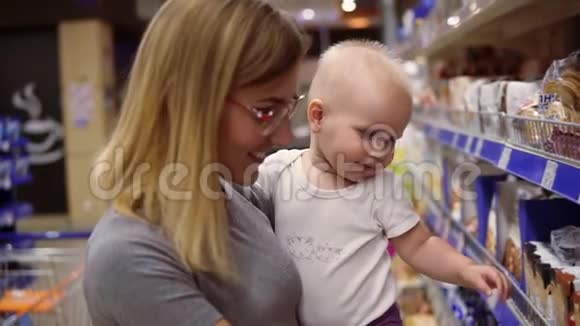 年轻的母亲戴着眼镜抱着孩子一边在超市的货架上挑选饼干视频的预览图