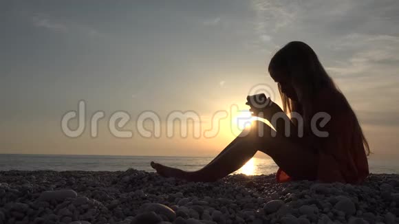 4K儿童在海滩上玩智能手机日落海海岸线上的女孩剪影视频的预览图