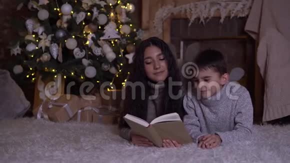 在圣诞节妈妈给儿子读了一本书吻他躺在圣诞树附近的地板上视频的预览图